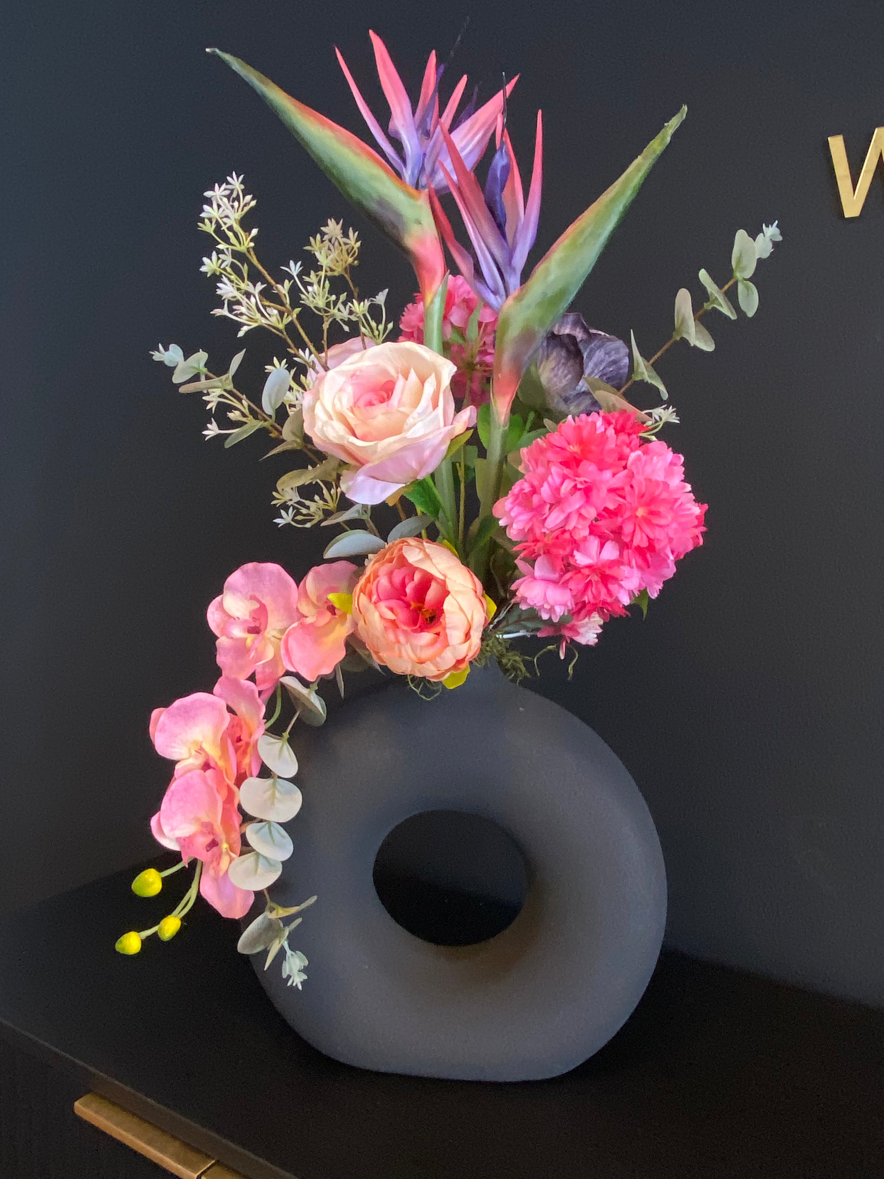 Tropisch luxe kunstbloemstuk met roze orchideeën, paradijsvogelbloem, peachkleurige bloemen en donkergrijze organische vaas.