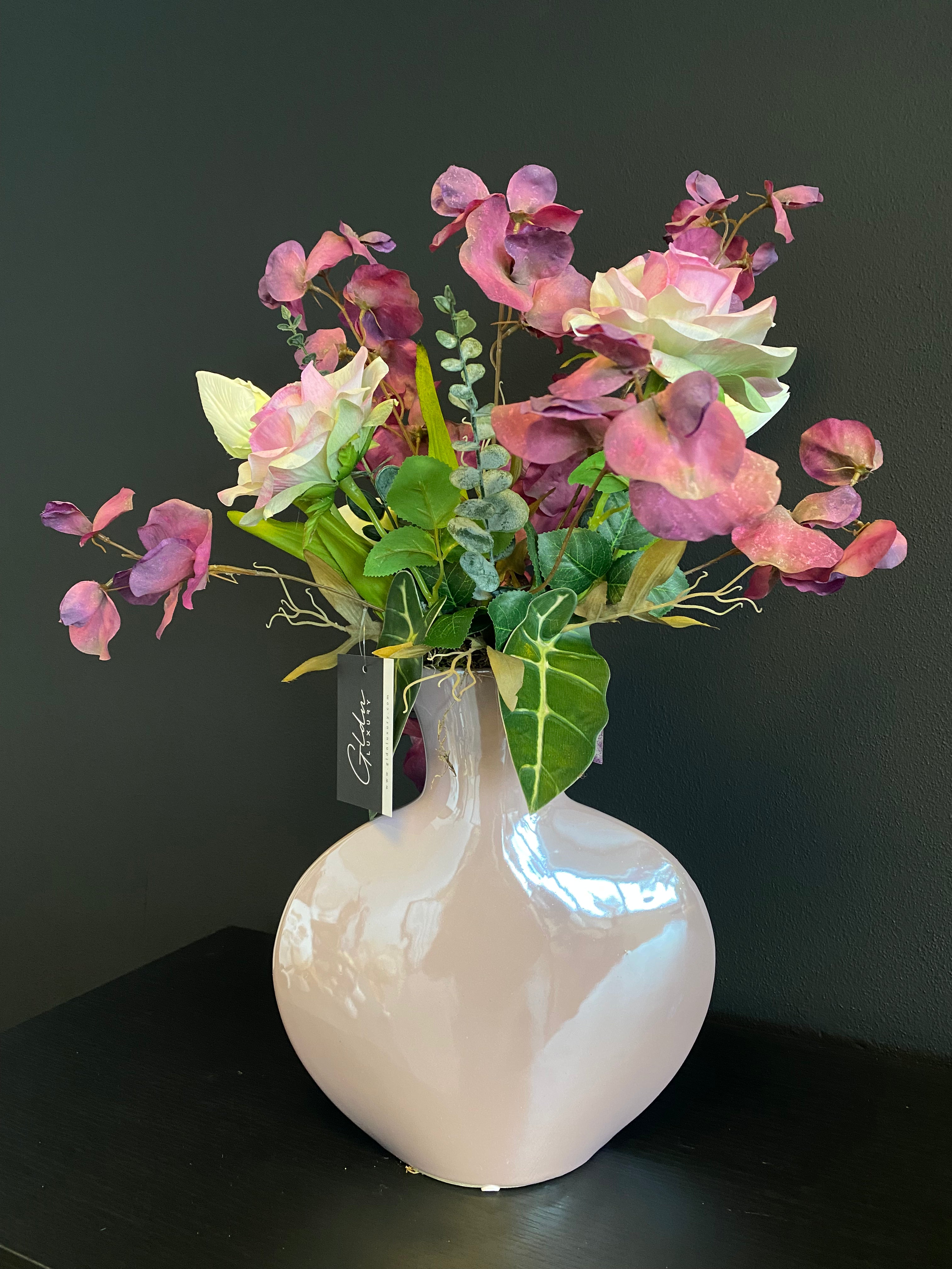 Luxe paars-roze kunstbloemstuk met roze, paarse en crèmekleurige bloemen in een glanzende roze designvaas van circa 50 cm hoog.